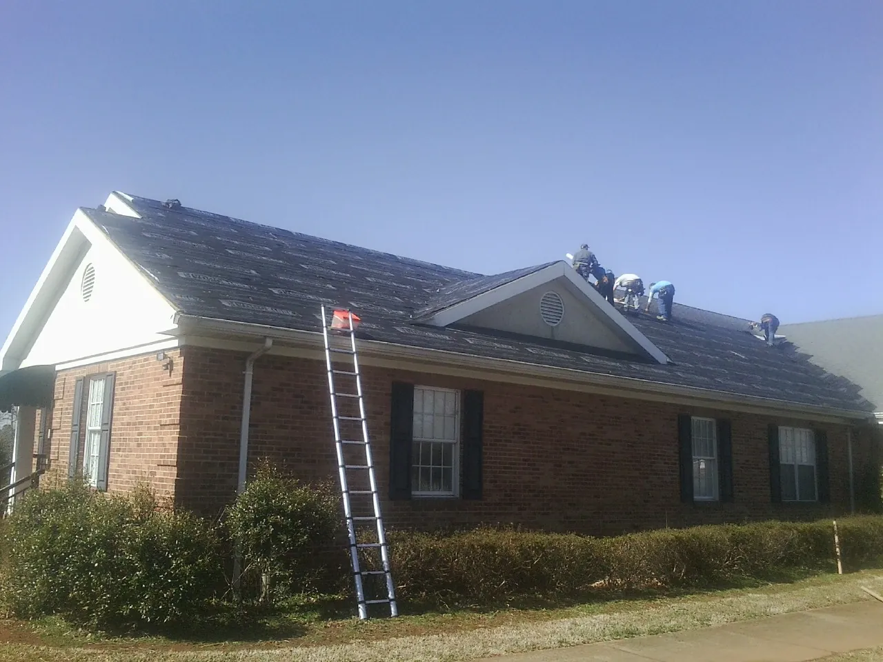 Crew tearing shingles off a brick ranch roof with a ladder set — Emergency Roof Repair work by our crew in Ellington