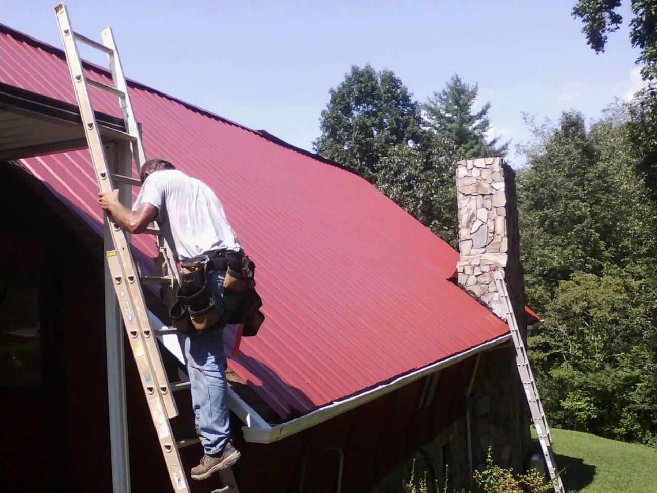 Roofer on a ladder beside a red metal roof and stone chimney — Soffit & Fascia Repair by Roofing Brothers in Ellington